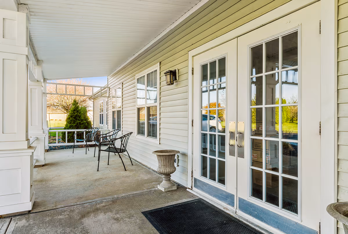 Covered outdoor porch area with white double glass doors, beige siding, two black metal chairs, a small stone planter, and a black doormat. Green grass and trees are visible in the background.