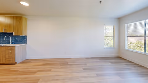 Empty open-plan living area with wood floors, a small kitchenette with wood cabinets and blue tile backsplash on the left, and large windows on the right.