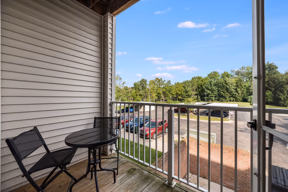 A small balcony with a round black table and two black chairs. The balcony has a white railing and overlooks a parking lot with several parked cars and green trees in the background under a blue sky with a few clouds.