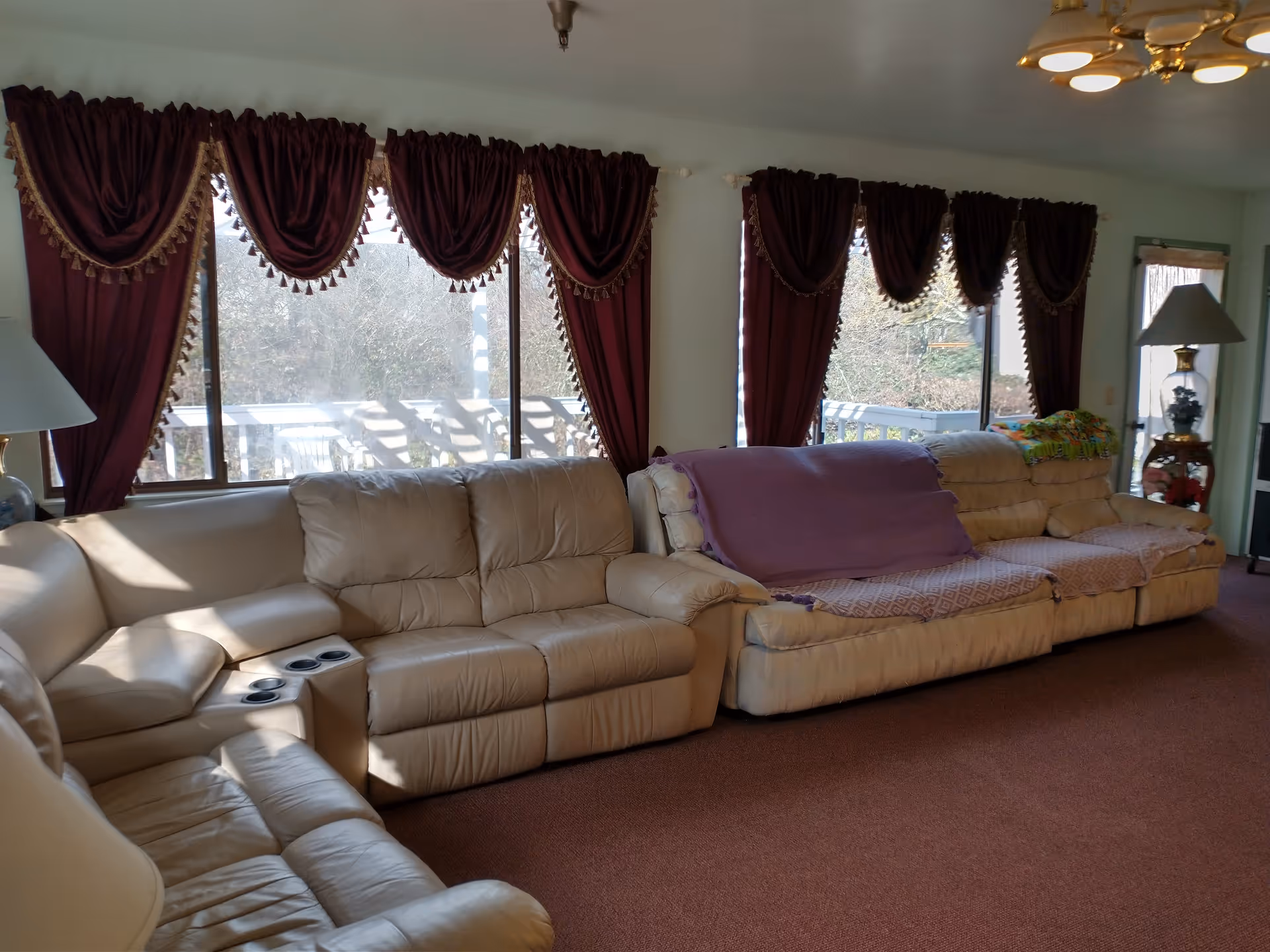 A living room with beige leather sofas arranged along the wall under windows with burgundy curtains. There are blankets draped over some of the sofas. A floor lamp with a white shade and a brass base is visible near a door with a window. The room has a brown carpet and a ceiling light fixture with multiple bulbs.