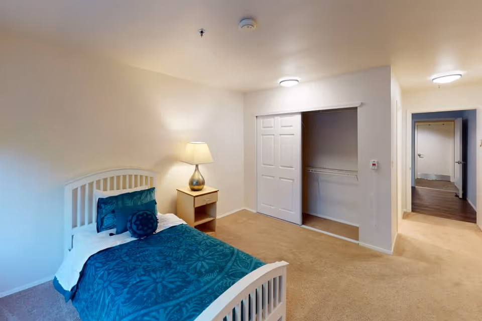 Small furnished bedroom with a single bed dressed in teal bedding, a nightstand and lamp, an open closet and a doorway to a hallway.