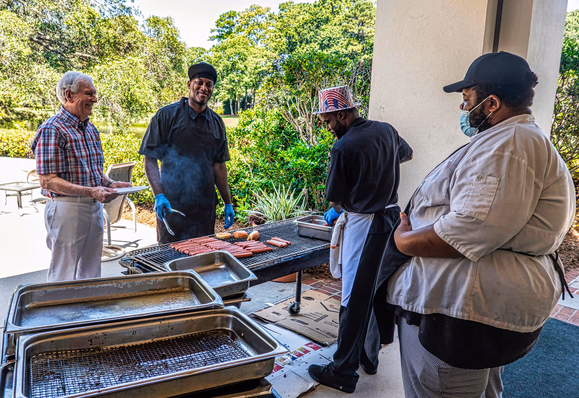 An elderly man in a plaid shirt holding a plate stands next to three men cooking hot dogs and hamburgers on a large outdoor grill. The men cooking are wearing black and white chef uniforms, with one wearing a patriotic hat and another wearing a face mask. The scene is set outdoors with green trees and bushes in the background.