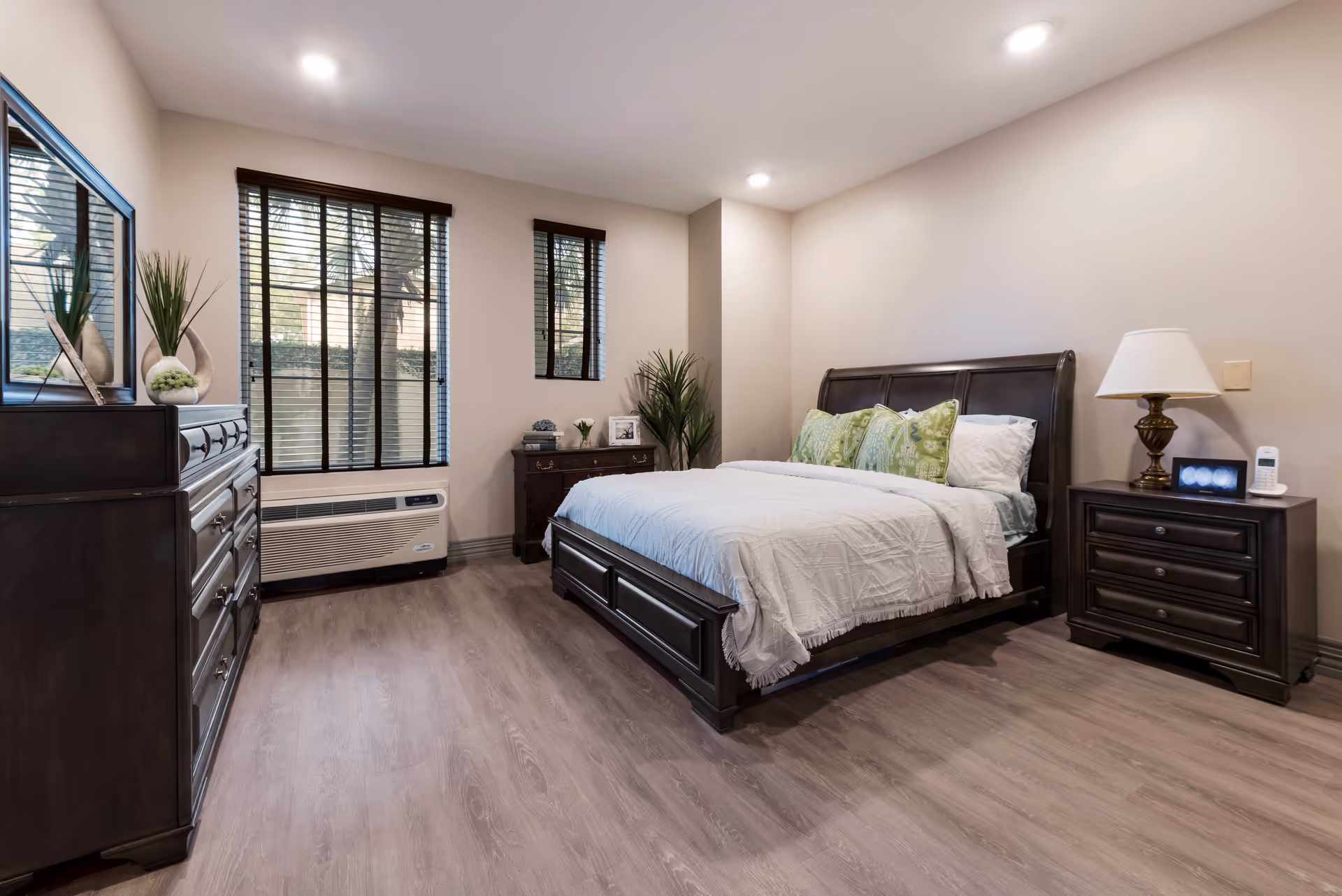 A neatly arranged bedroom with a large dark wooden bed featuring white bedding and green decorative pillows. The room has light-colored walls and wood flooring. There is a matching dark wooden dresser with a mirror on the left side, a nightstand with a lamp and a phone on the right side, and a small chest of drawers with decorative items and plants near the window. The window has dark blinds, and there is a wall-mounted air conditioning unit below it.