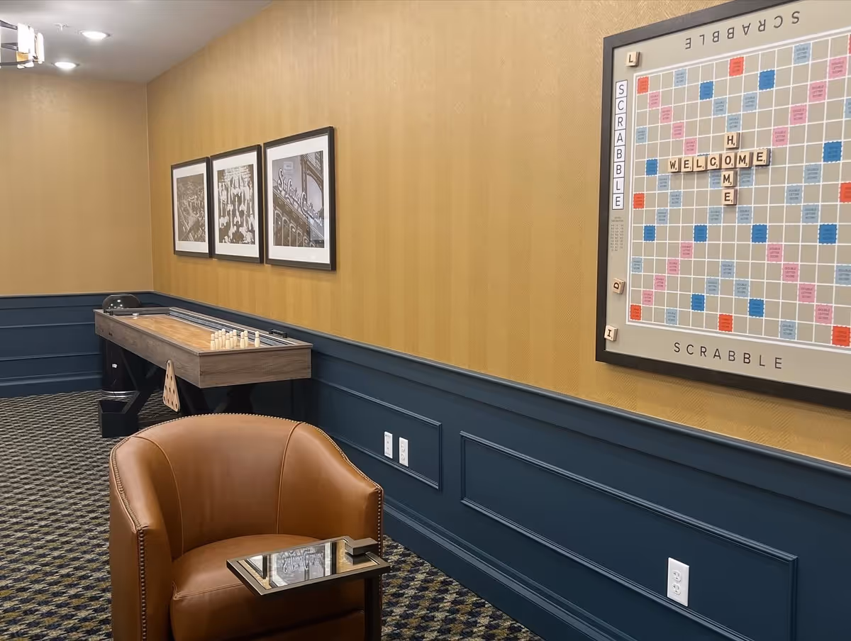 A cozy game room with a brown leather armchair and a small glass side table in the foreground. On the wall, there is a large framed Scrabble board with the words 'WELCOME HOME' spelled out. The walls are painted mustard yellow on the upper half and dark blue on the lower half with decorative molding. There is a shuffleboard table along the wall with small pins set up, and three black and white framed photographs hang above it. The carpet has a patterned design in dark colors.