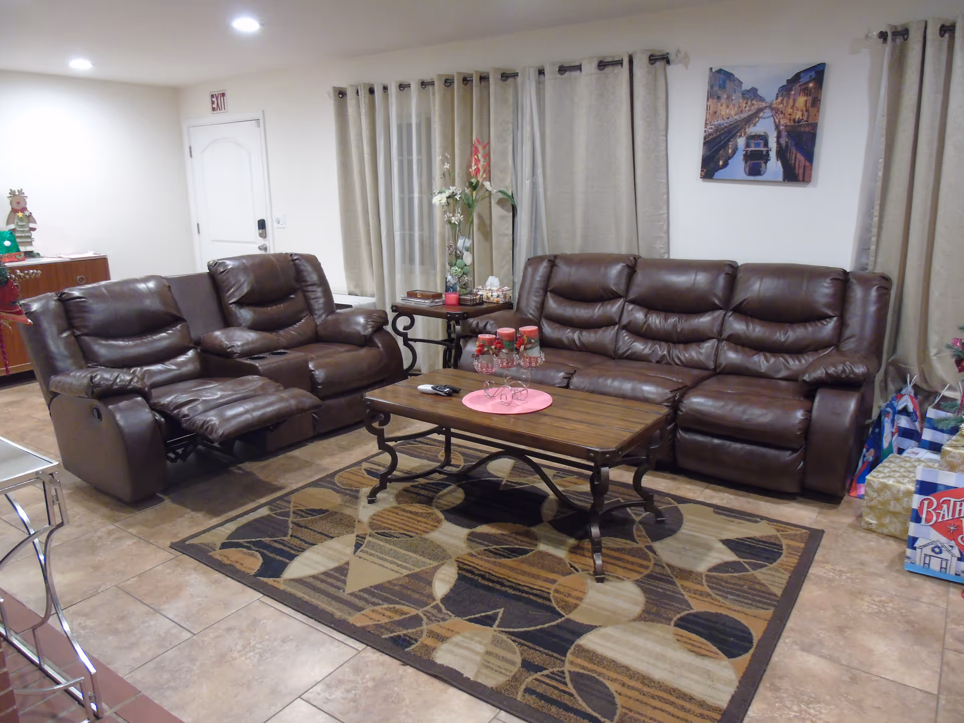 A cozy living room with two brown leather reclining sofas facing a wooden coffee table on a patterned area rug. The room has beige tiled flooring, beige curtains covering windows, a side table with decorative items, and a wall picture of a canal scene. There are some gift bags and wrapped presents in the corner.