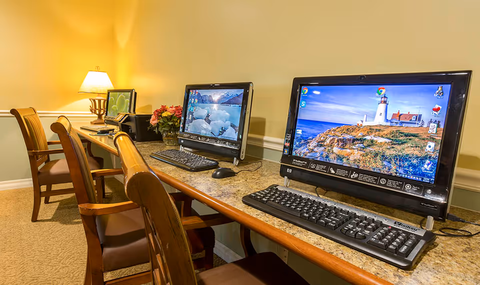 A row of three desktop computers on a long counter with wooden chairs in front of each. The room has beige walls and carpet, with a table lamp and a vase of flowers on the counter.