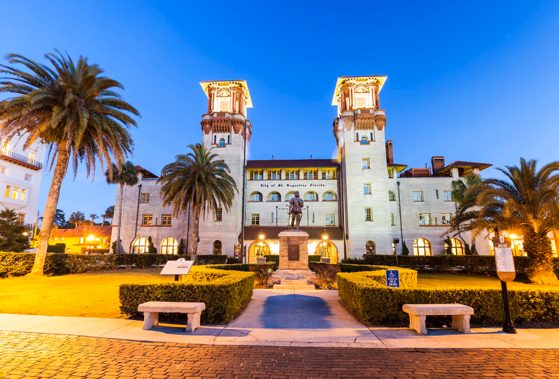Evening view of a historic building with two tall towers, palm trees, and a statue in front. The building is illuminated with warm lights and has a sign that reads 'City of St. Augustine Florida'. There are benches and trimmed hedges along the walkway leading to the statue.