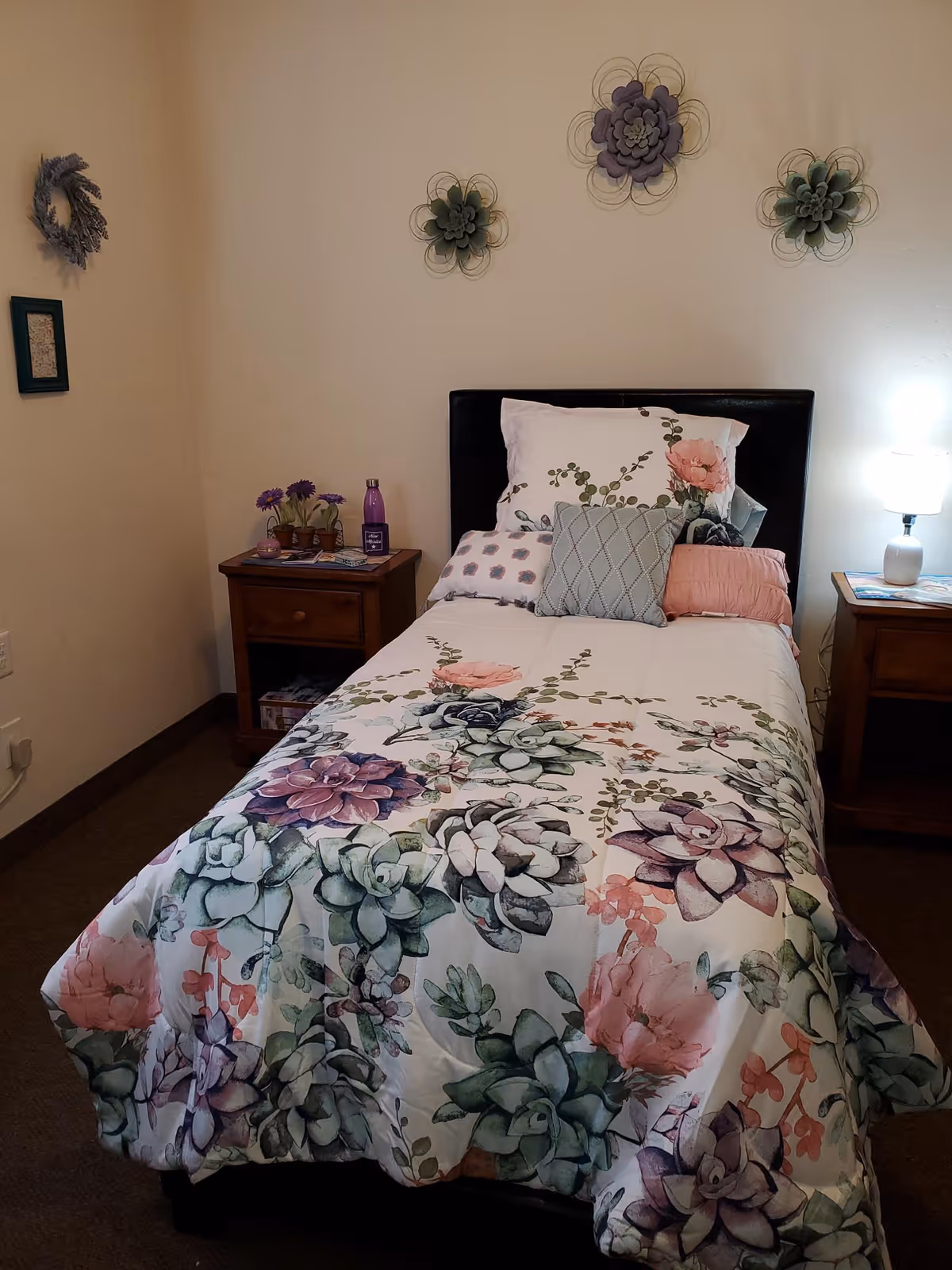 A cozy bedroom with a single bed featuring a floral patterned comforter and multiple pillows. The bed has a dark headboard and is flanked by two wooden nightstands. One nightstand has a small lamp turned on, and the other has a purple water bottle and some small decorative items. The wall above the bed is decorated with three metal flower wall hangings.