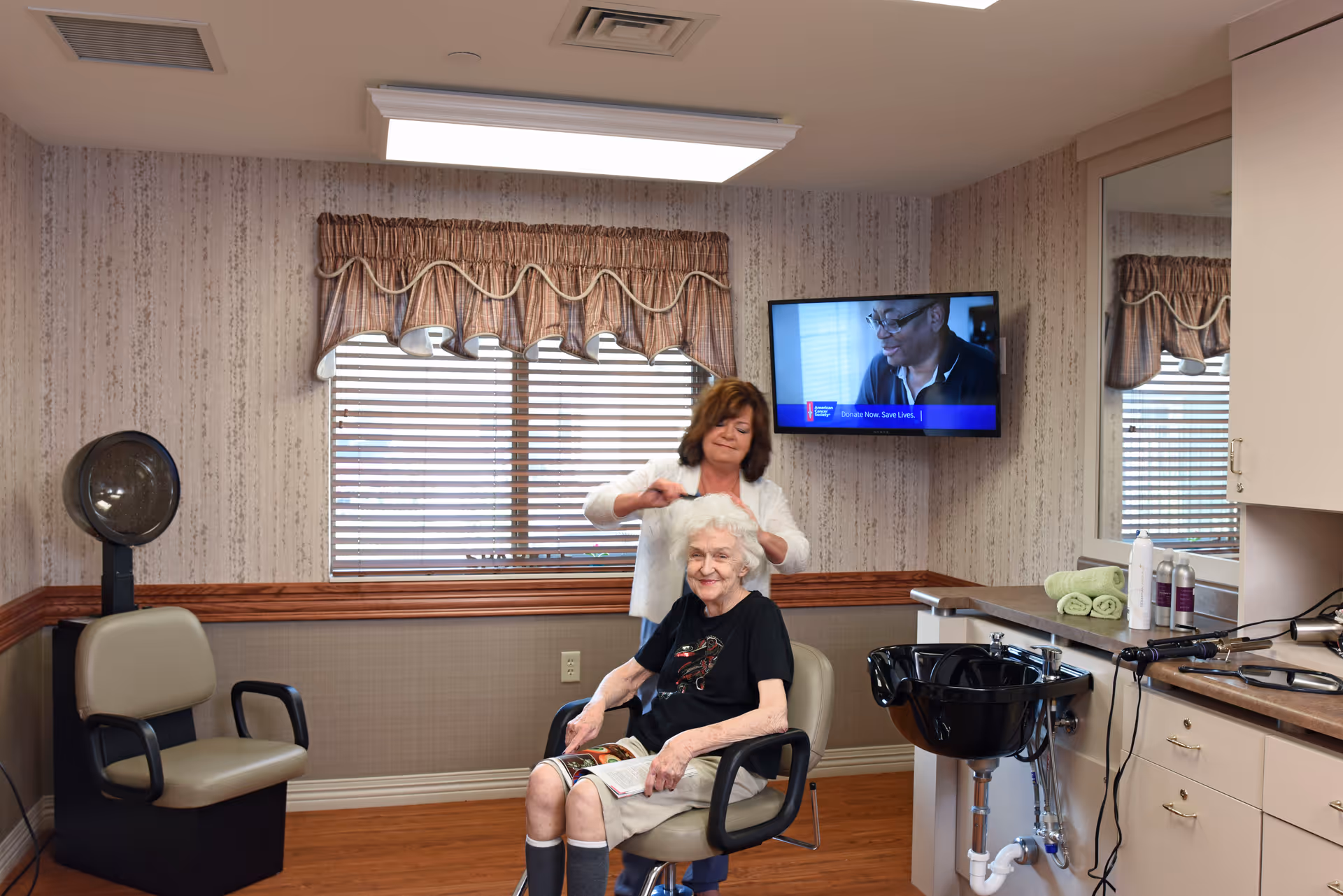 An elderly woman sitting in a salon chair reading a magazine while a hairstylist stands behind her, styling her white hair. The room has wood flooring, a window with blinds and a valance, a mounted television on the wall, and salon equipment including a black hair washing sink and hair care products on the counter.
