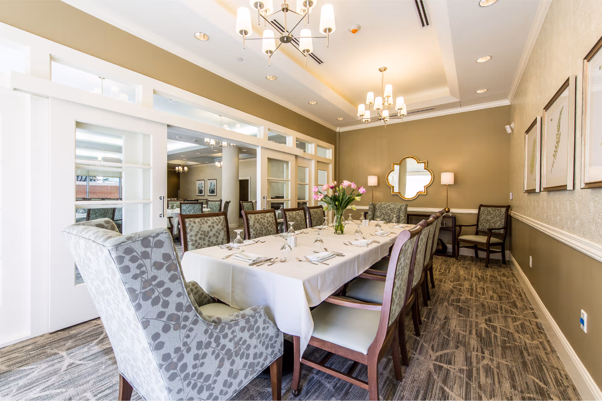 A well-lit dining room with a long rectangular table covered with a white tablecloth, set with glassware, napkins, and silverware. The table is surrounded by cushioned chairs with patterned upholstery. A vase with pink tulips is placed in the center of the table. The room features beige walls, a decorative mirror, framed botanical prints, and modern chandeliers hanging from the ceiling. Sliding glass doors separate this room from another dining area visible in the background.