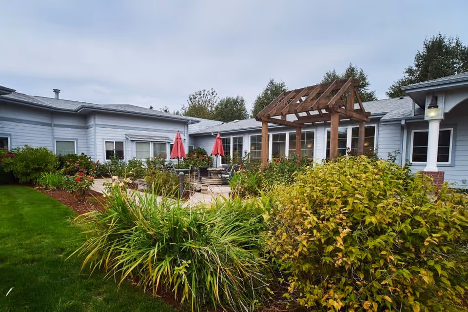 Outdoor patio area at Sweetbriar Villa featuring a wooden pergola, green shrubs, and plants with a building in the background. There are red umbrellas and seating arrangements on the patio.