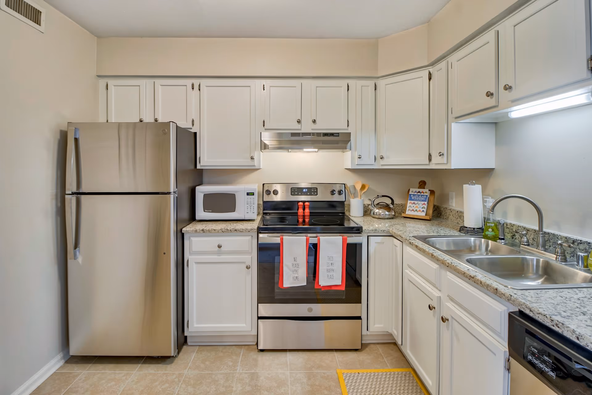 A clean and modern kitchen with white cabinets, granite countertops, and stainless steel appliances including a refrigerator, stove, microwave, and dishwasher. Two red and white kitchen towels hang on the oven door, and various kitchen utensils and a kettle are placed on the countertops.