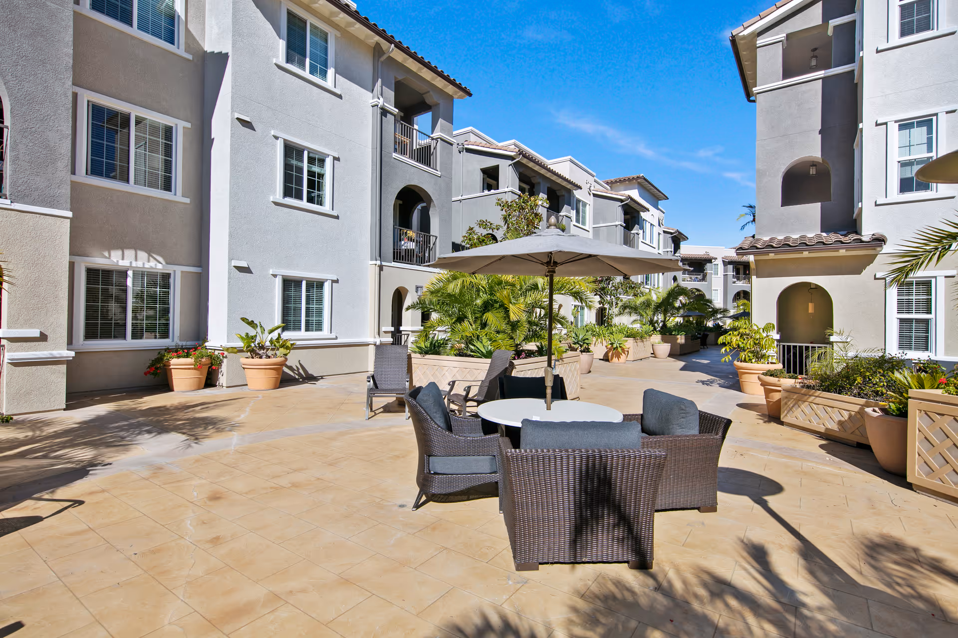 Outdoor courtyard area at Meridian at Lake San Marcos featuring a round table with four wicker chairs and a large umbrella. The courtyard is surrounded by multi-story residential buildings with balconies and windows, potted plants, and clear blue sky overhead.