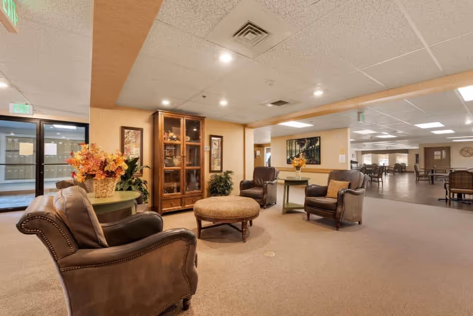 A spacious and well-lit common area in a senior living facility featuring four brown leather armchairs arranged around a round upholstered ottoman. There are two small tables with flower arrangements, a wooden display cabinet against the wall, and framed artwork. The area extends into a dining space with tables and chairs visible in the background.