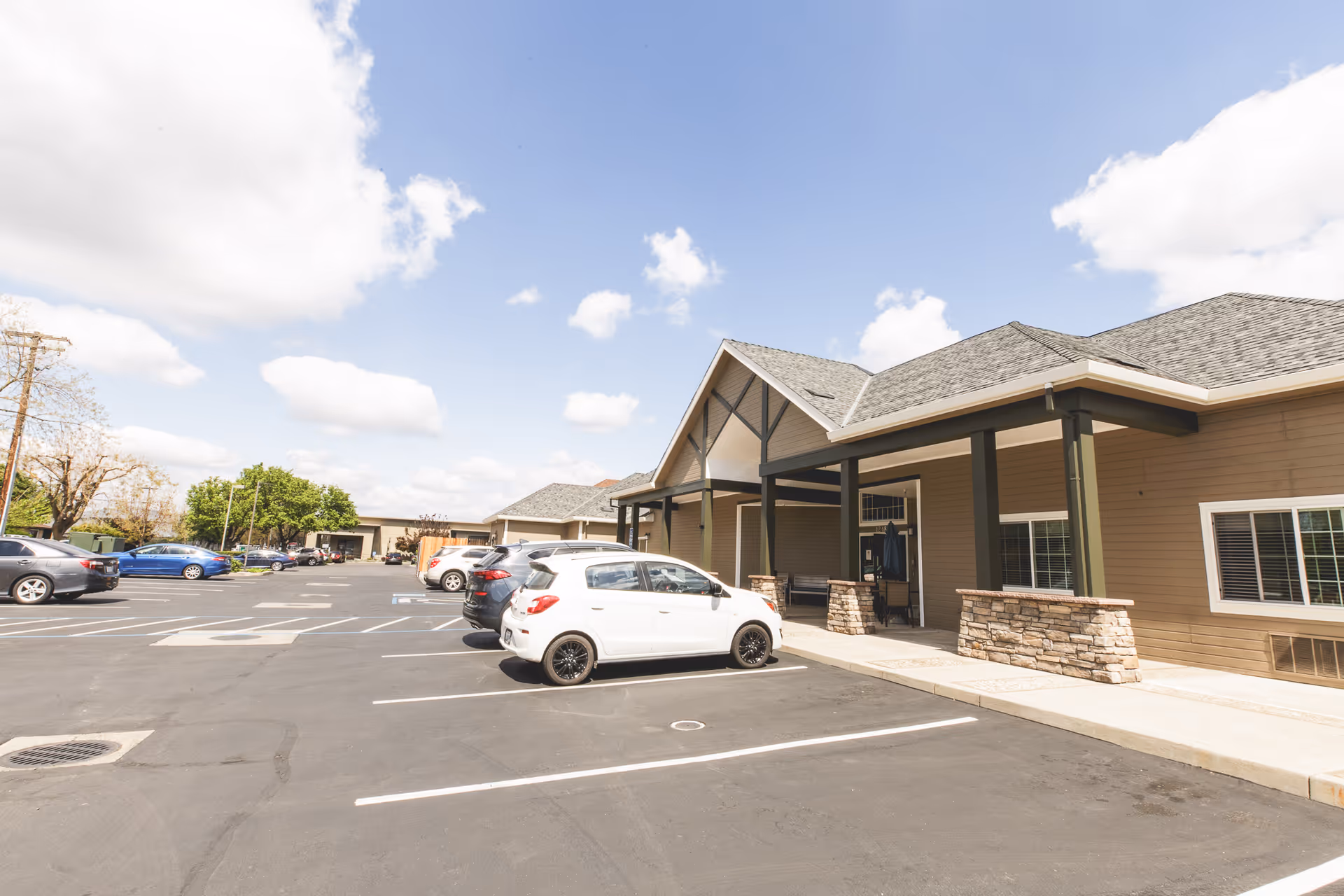 Exterior view of Orangeburg Manor showing a single-story building with a covered entrance supported by stone pillars. Several cars are parked in the parking lot in front of the building under a partly cloudy sky.