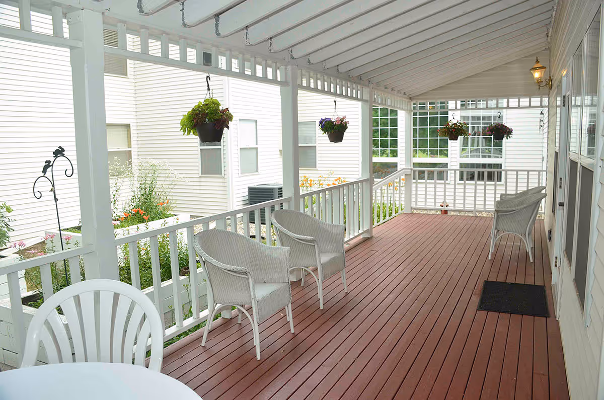 Covered porch area with white railing and ceiling, featuring several white wicker chairs and a white plastic chair around a round table. Hanging flower pots with green and purple plants are suspended from the ceiling. The porch overlooks a garden area with flowers and is adjacent to a white building.