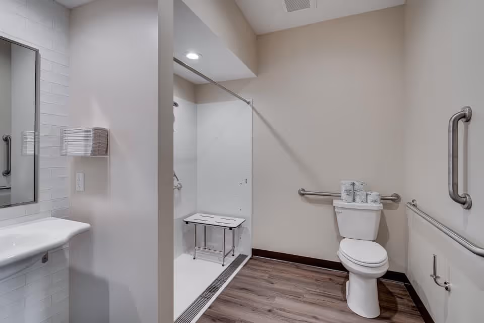 A senior living facility bathroom featuring a walk-in shower with a foldable bench and grab bars, a white toilet with several rolls of toilet paper on top, a wall-mounted sink, a mirror, and grab bars on the walls. The floor has wood-like vinyl flooring and the walls are painted beige.