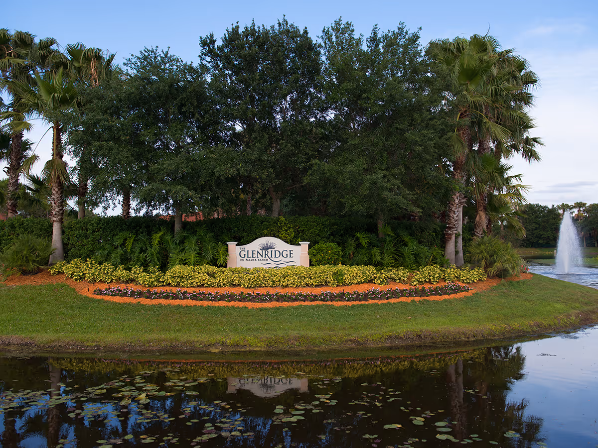 Landscaped entrance with a stone sign reading "The Glenridge on Palmer Ranch" surrounded by palm trees, flower beds, a pond, and a fountain.