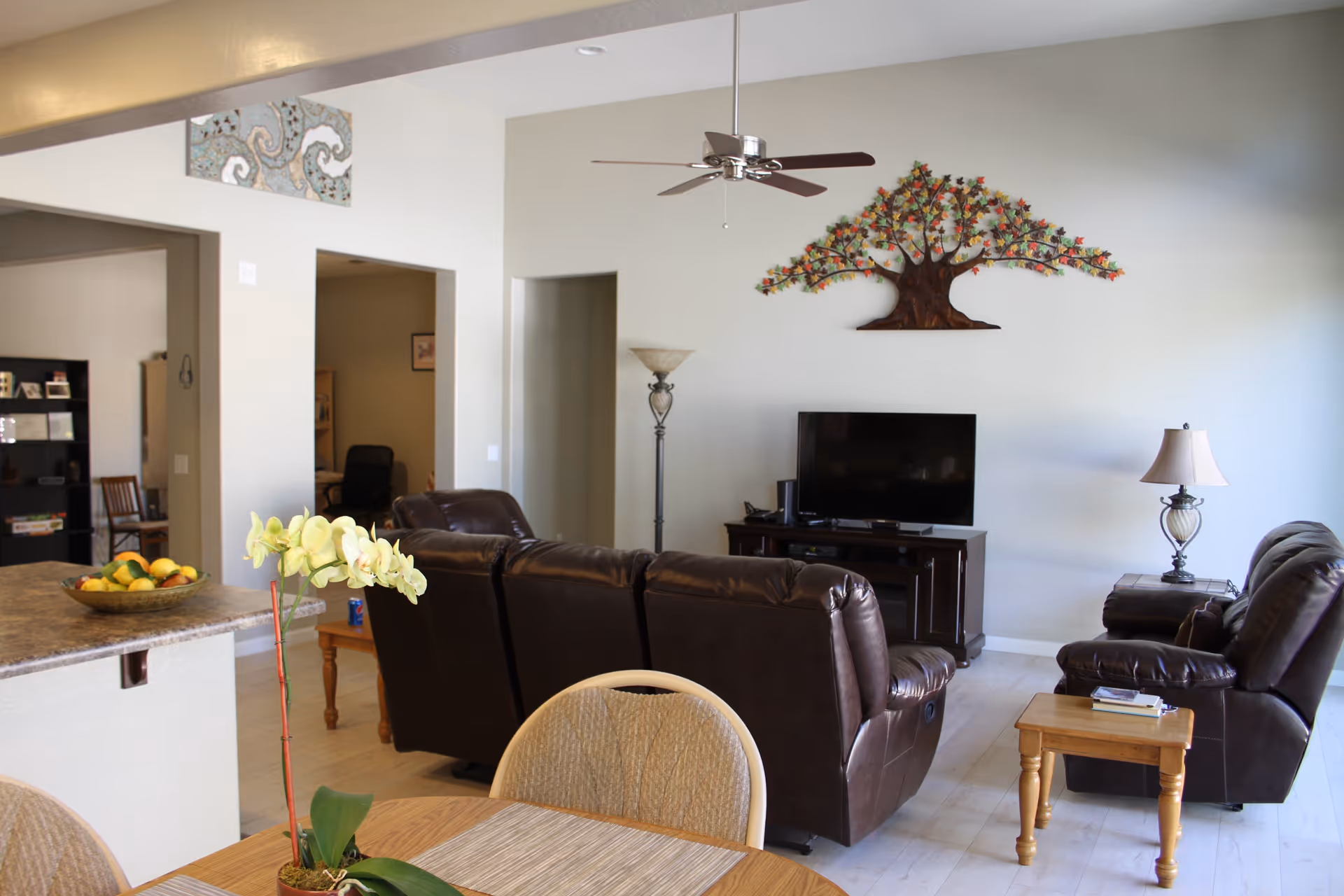 A bright living room with a brown leather sofa set facing a flat-screen TV on a wooden stand. There is a decorative metal tree wall art above the TV. A ceiling fan with wooden blades hangs from the ceiling. To the left, a kitchen counter with a bowl of fruit and a yellow orchid plant on a dining table with beige chairs are visible. The room has light-colored walls and flooring, with a floor lamp and a table lamp providing additional lighting.