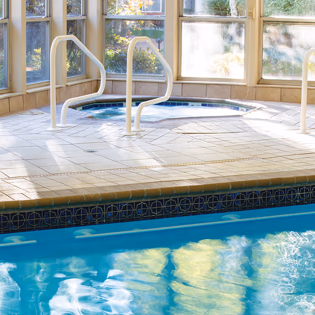 Indoor swimming pool with an adjacent raised hot tub, handrails, tiled deck and large windows.