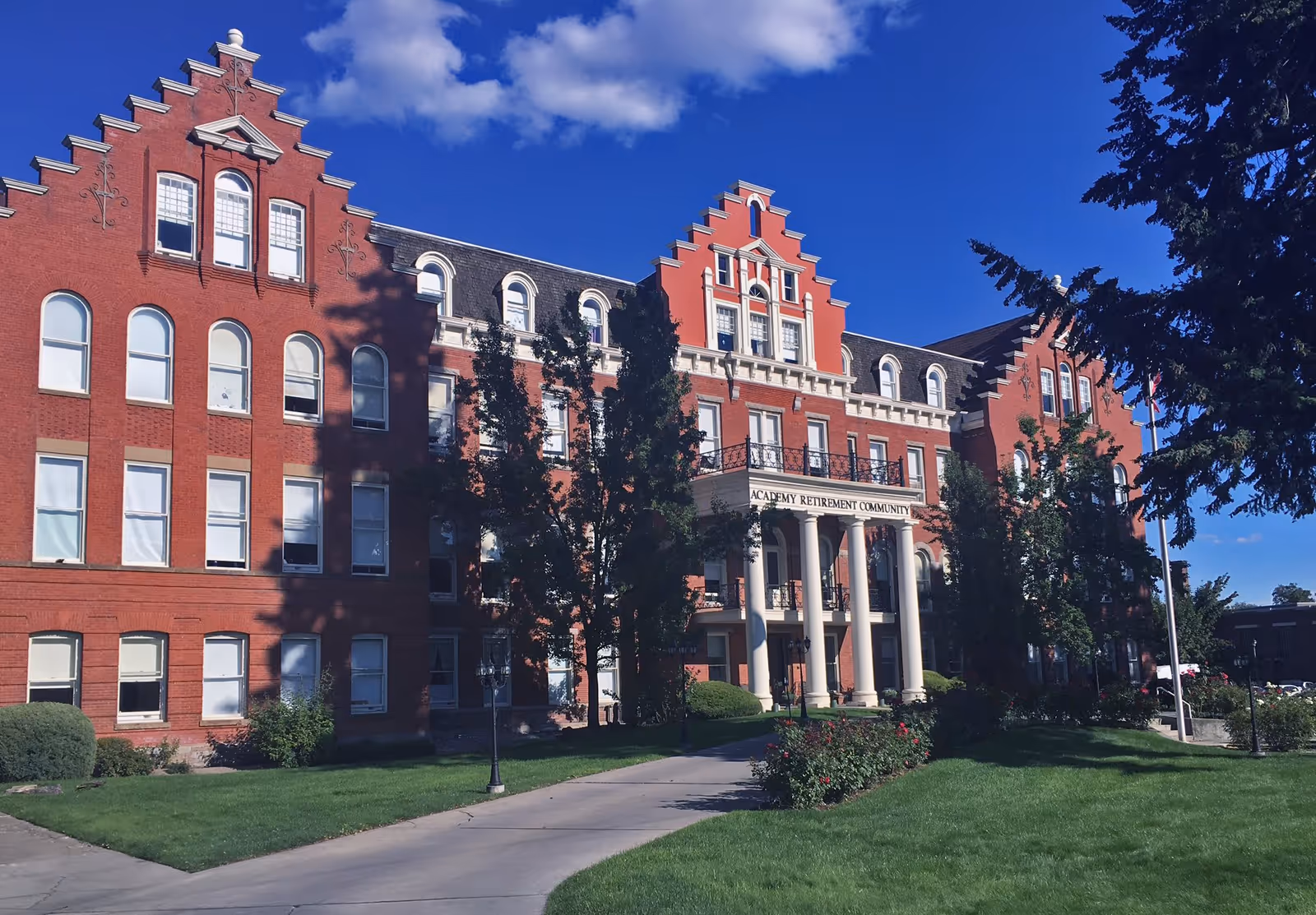 Exterior view of a large red brick building with multiple windows and decorative gables under a blue sky with some clouds. The building has a prominent entrance with white columns and a sign that reads 'Academy Retirement Community'. There are trees and green lawns in front of the building.