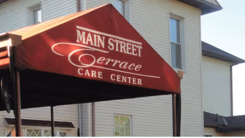 Red canopy with white text reading 'Main Street Terrace Care Center' attached to the exterior of a beige multi-story building with several windows.
