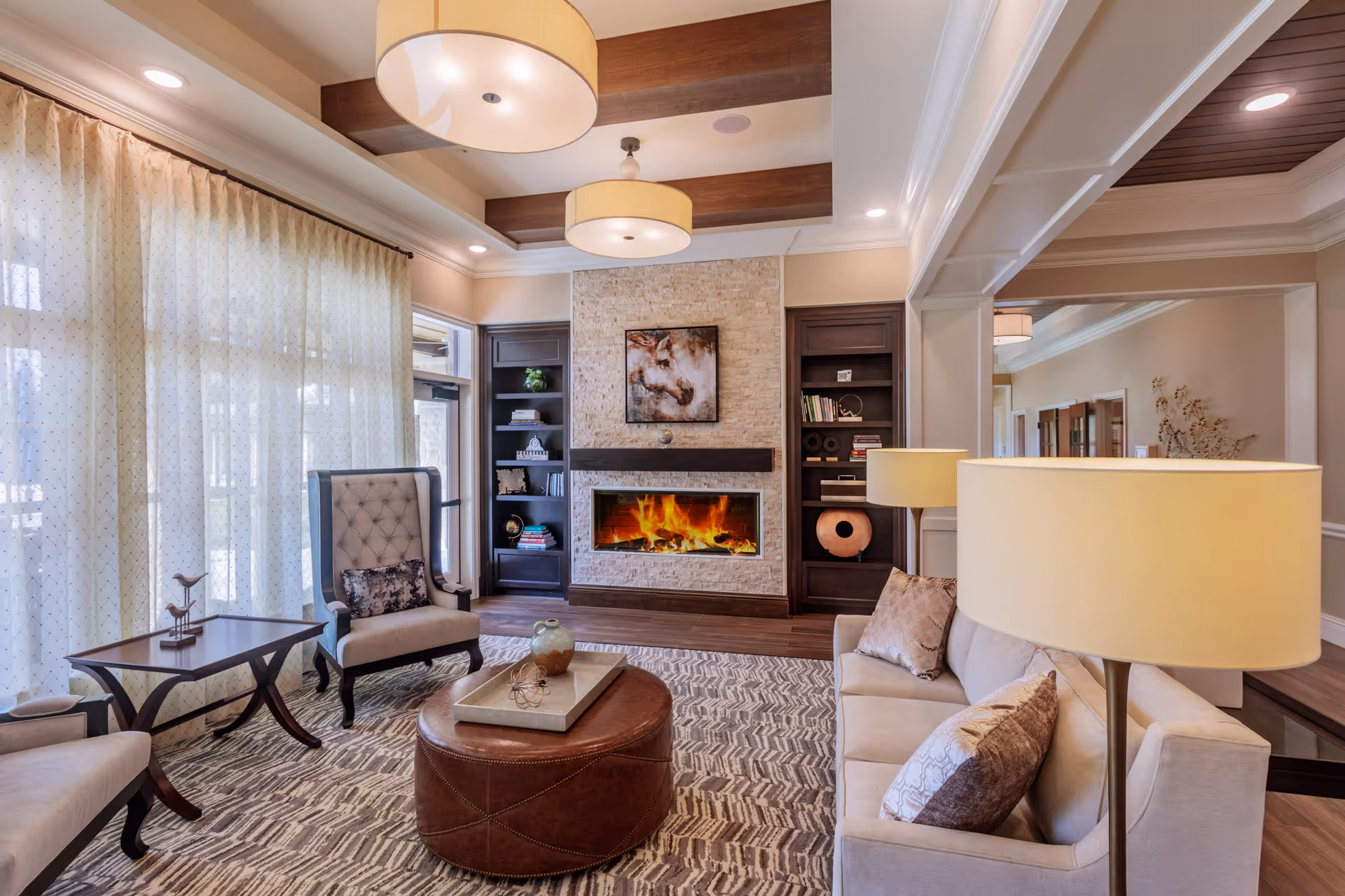 A cozy living room with a modern fireplace set in a stone wall flanked by dark wooden shelves filled with books and decorative items. The room features a beige sofa with pillows, a tufted armchair, a round leather ottoman with a tray on top, a side table, and two floor lamps with beige lampshades. Large windows with sheer curtains allow natural light to fill the space. The ceiling has wooden beams and two large round pendant lights.