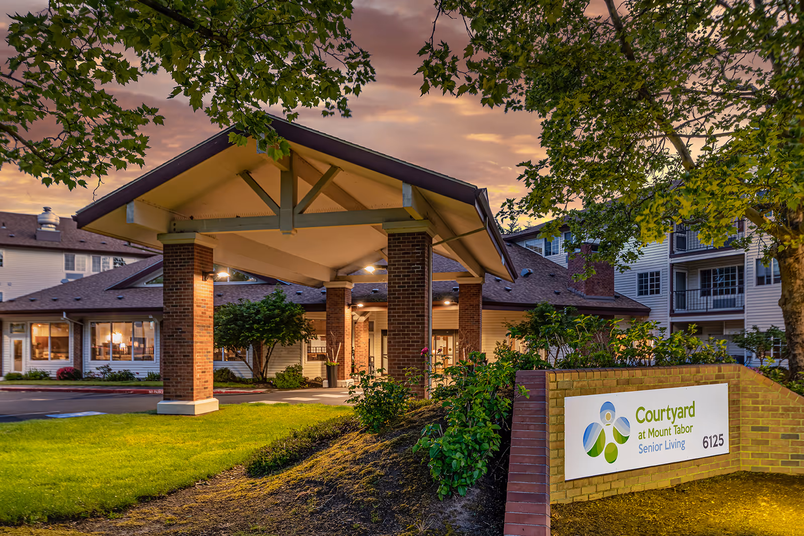 Exterior view of Courtyard at Mount Tabor senior living facility at dusk, showing the entrance canopy supported by brick pillars, well-maintained landscaping, and a sign with the facility name and address.