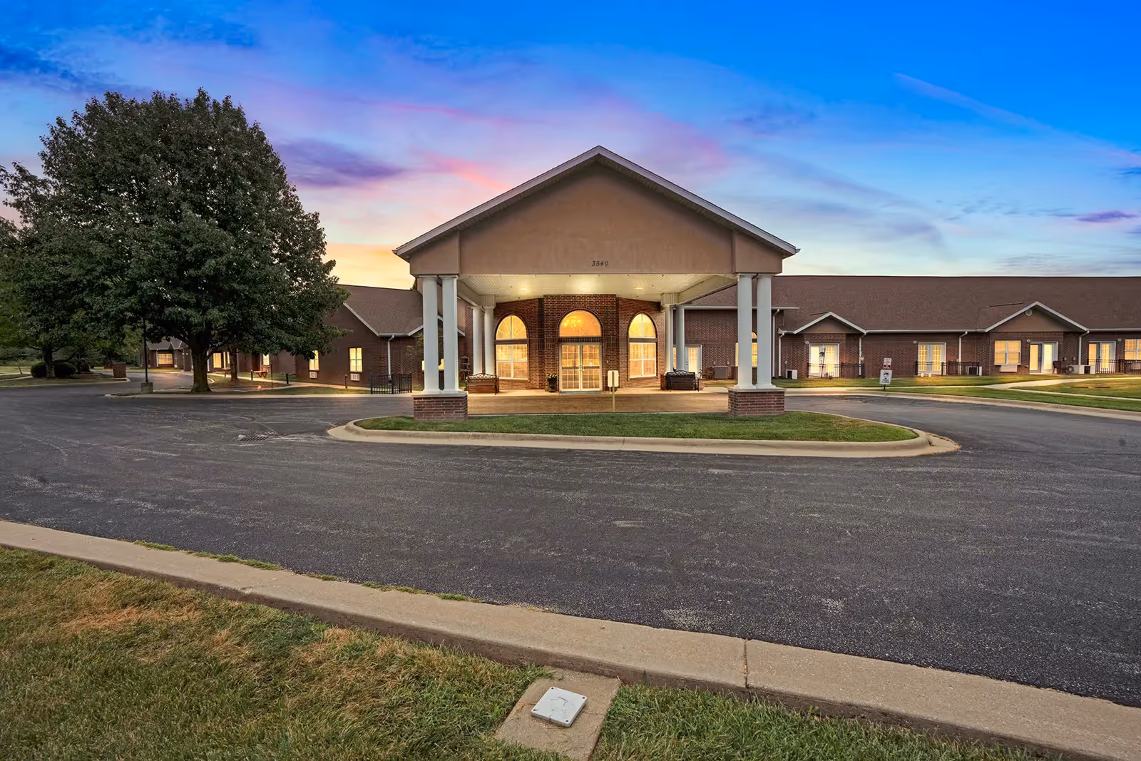 Front entrance of a single-story senior living building with a covered porte-cochere, lit windows, and a circular driveway at sunset.