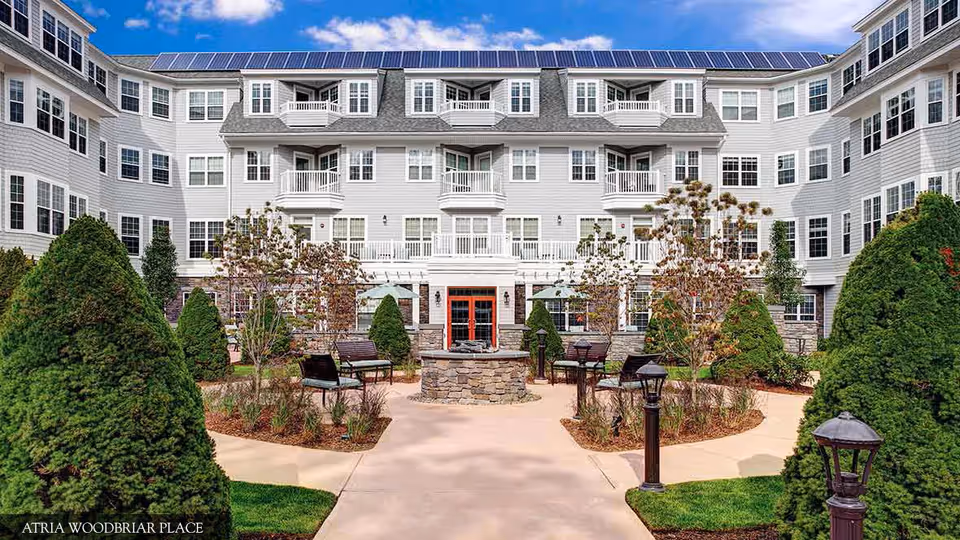 Exterior view of a senior living facility with a courtyard featuring a circular stone fire pit, benches, landscaped bushes, and trees. The building is multi-story with numerous windows, balconies, and solar panels on the roof under a partly cloudy blue sky.