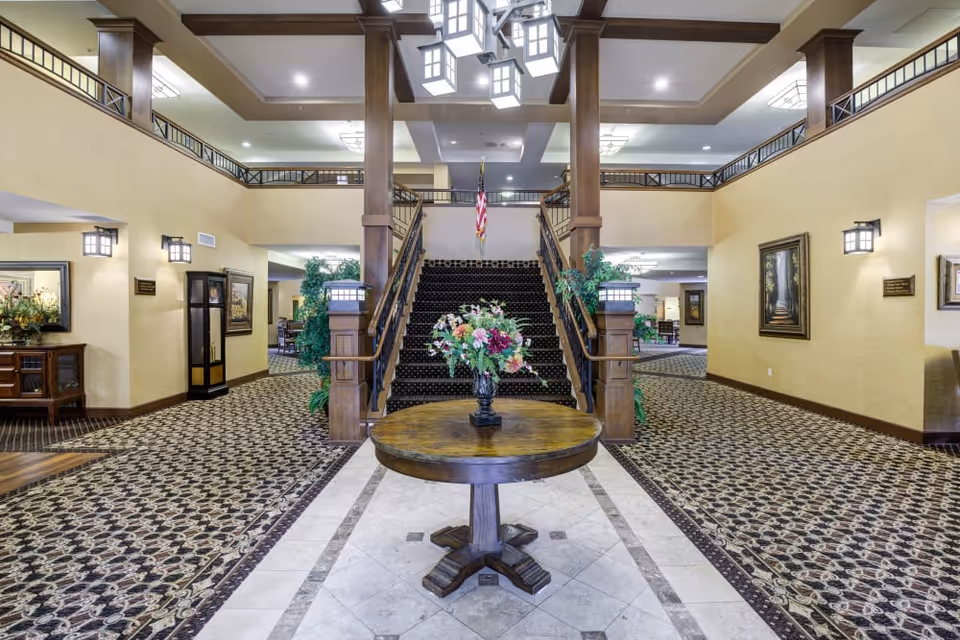 Spacious two-story lobby with a central round table topped by a floral arrangement and a staircase leading to an upper mezzanine.