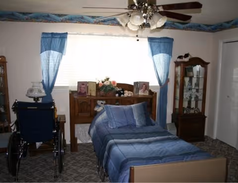 Blue-themed bedroom in a senior care home with a bed, wheelchair, and dresser.