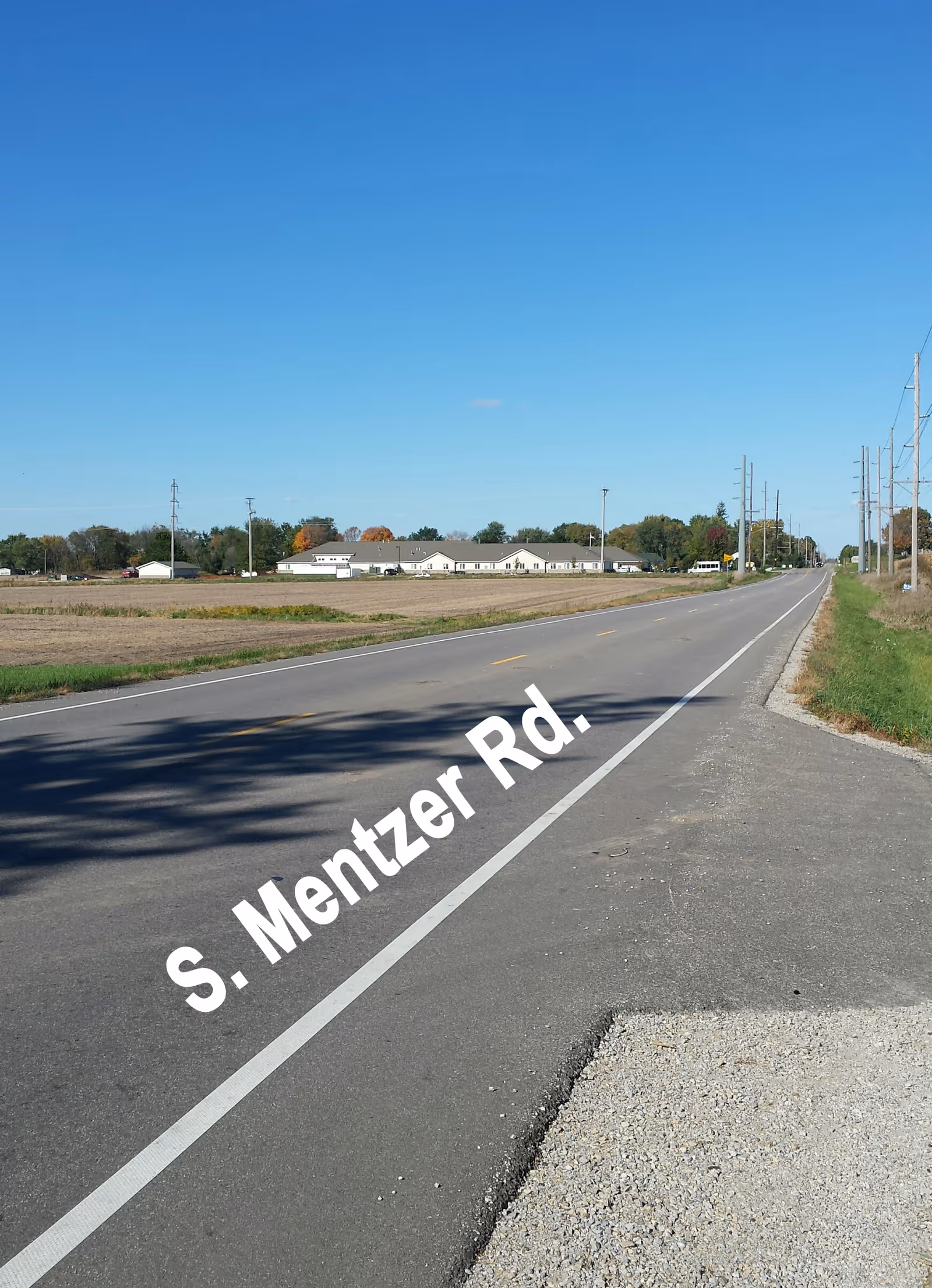 A long, straight rural road named S. Mentzer Rd. stretches into the distance under a clear blue sky. On the left side of the road, there are open fields and a single-story building in the background surrounded by trees. Utility poles line the right side of the road.