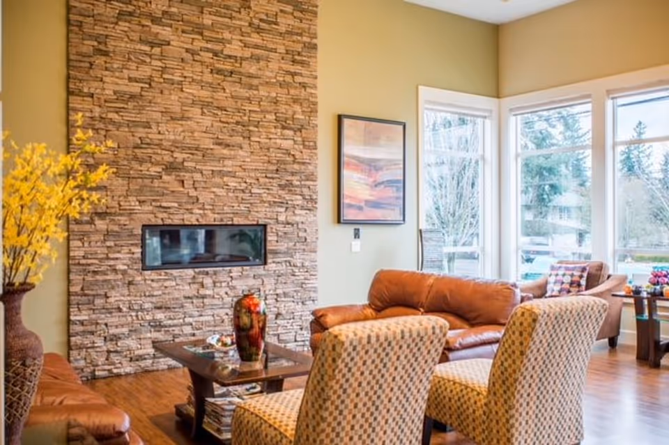 Sunlit living room with a stacked-stone fireplace, leather sofa, patterned armchairs, coffee table, and large windows.