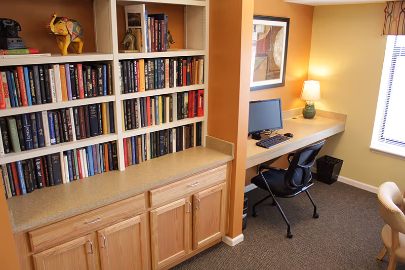 Interior view of a room with a built-in bookshelf filled with books and decorative items on the left. To the right, there is a desk with a computer, keyboard, mouse, and a lamp. A black office chair is positioned at the desk, and a window with blinds is visible on the far right.