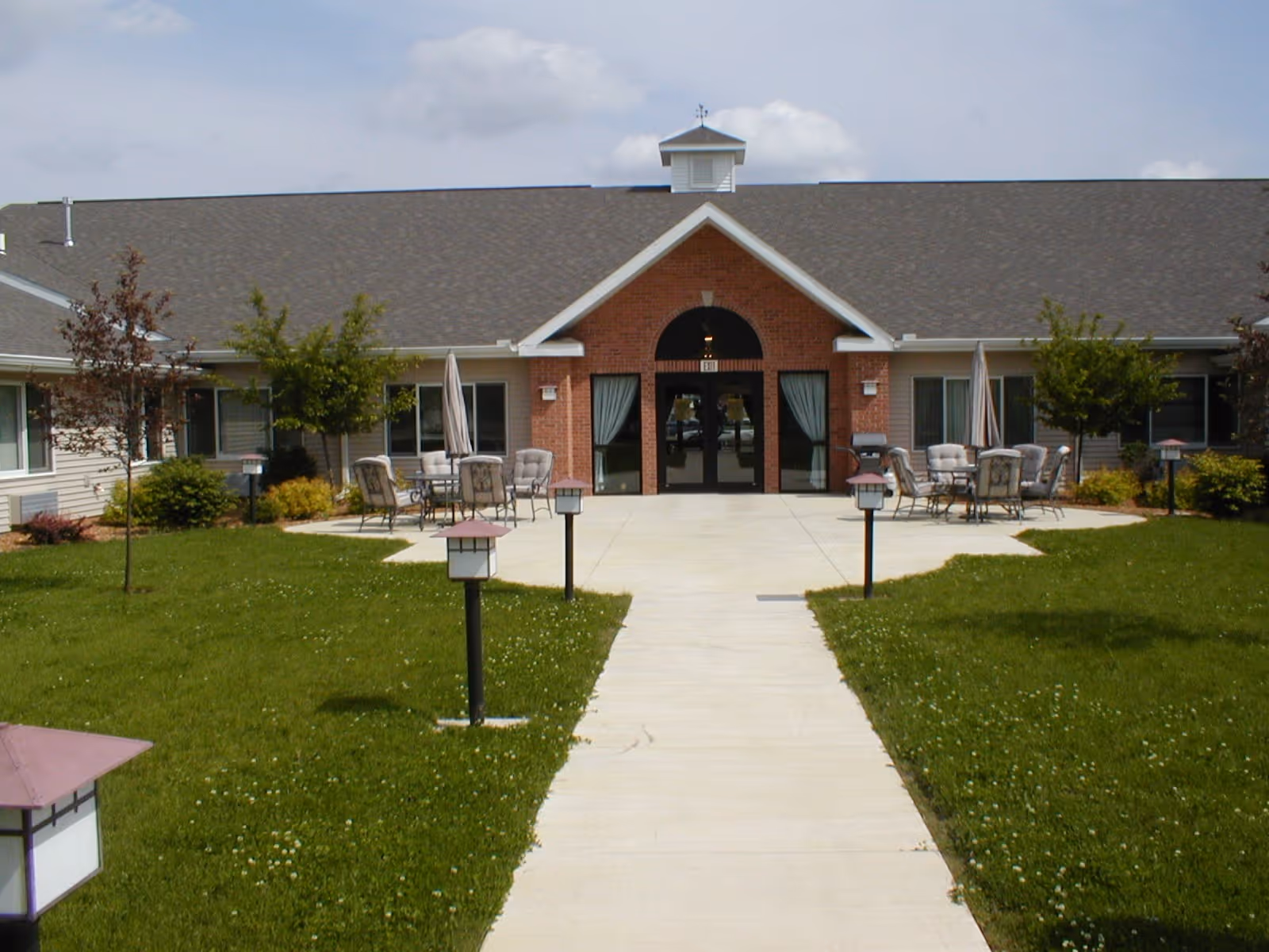 Walkway leading to the front entrance of a single-story brick and siding senior living building with outdoor seating and lawn.
