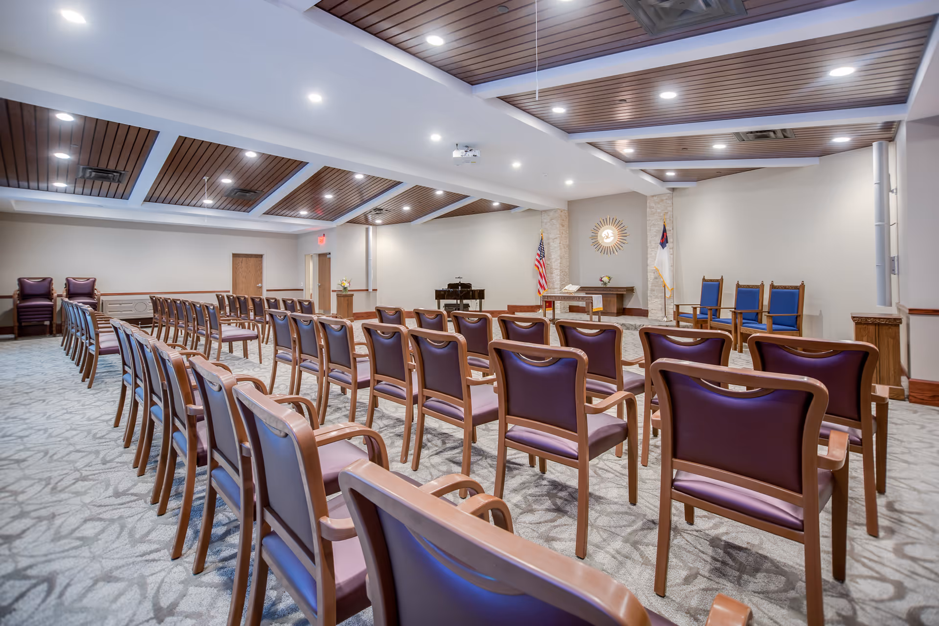 A spacious chapel or meeting room with rows of wooden chairs with purple cushions arranged facing a small altar area. The altar area has a table with an open book, a piano, and flags on either side. The ceiling features wooden paneling with recessed lighting.
