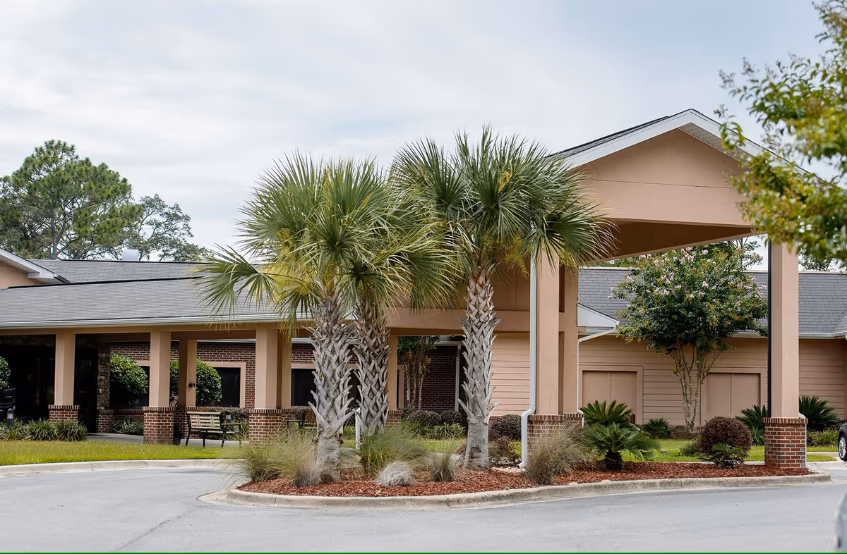 Front entrance of a single-story senior living building with a covered porte-cochère, palm trees, and a circular driveway.