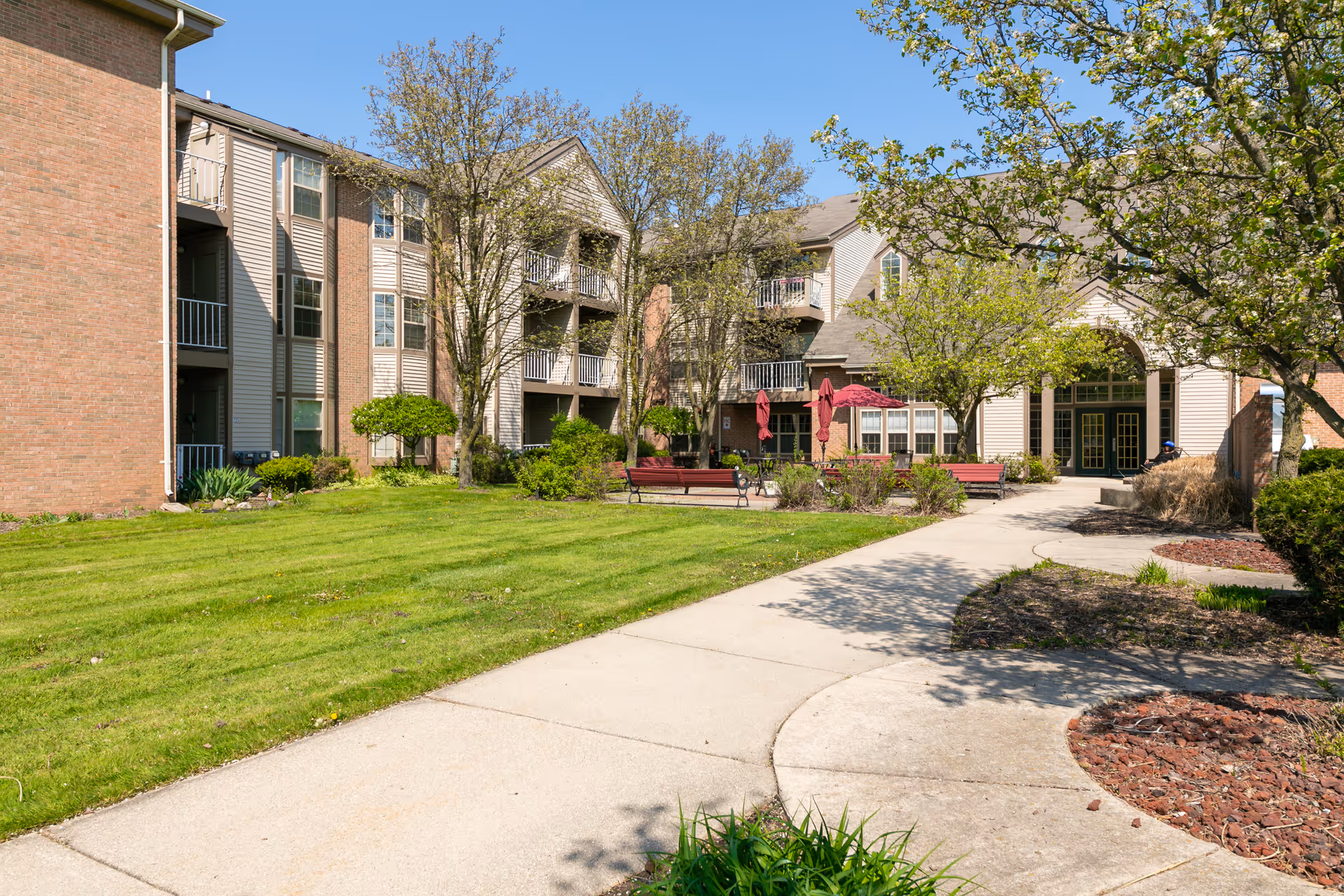 Courtyard and front facade of a three-story senior living building with a paved walkway, benches, umbrellas, and landscaped lawn.