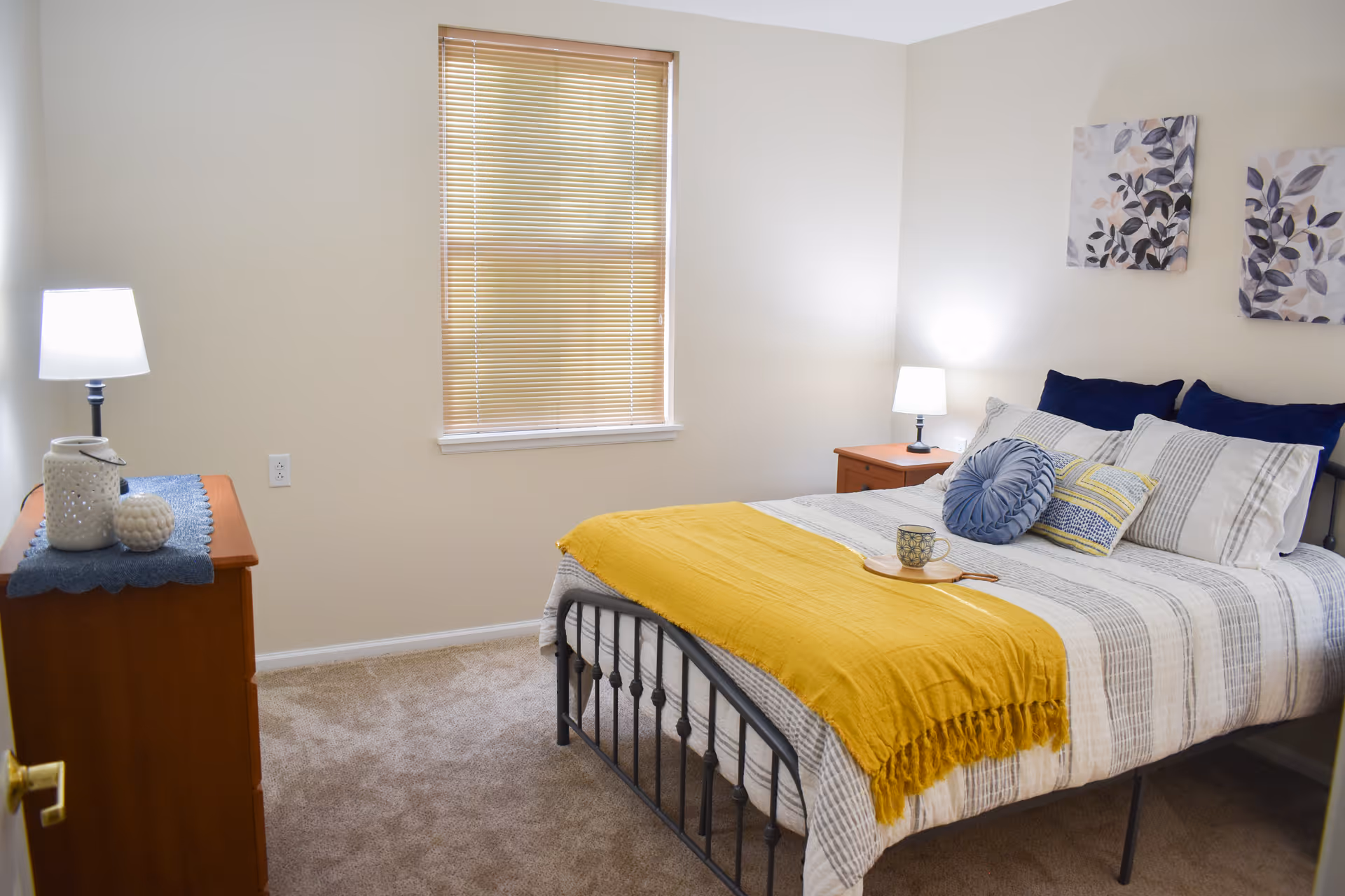 A cozy bedroom with a neatly made bed featuring striped bedding, a yellow throw blanket, and multiple decorative pillows. There are two wooden nightstands on either side of the bed, each with a lamp. A wooden dresser with decorative items and a lamp is positioned against the left wall. A window with closed blinds is centered on the back wall, and two framed leaf-themed artworks hang above the bed.