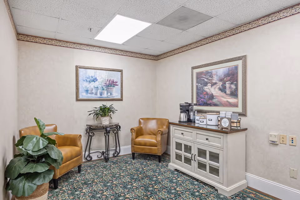 A cozy seating nook with two tan leather chairs, a small plant table, a cabinet coffee station, and framed artwork on the walls.