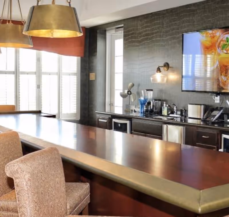 A modern kitchen area with a long countertop and upholstered chairs. The kitchen features dark cabinetry, a blender, a toaster, a small fan, and a mounted flat-screen TV displaying a drink. Large windows with white shutters allow natural light to fill the space.