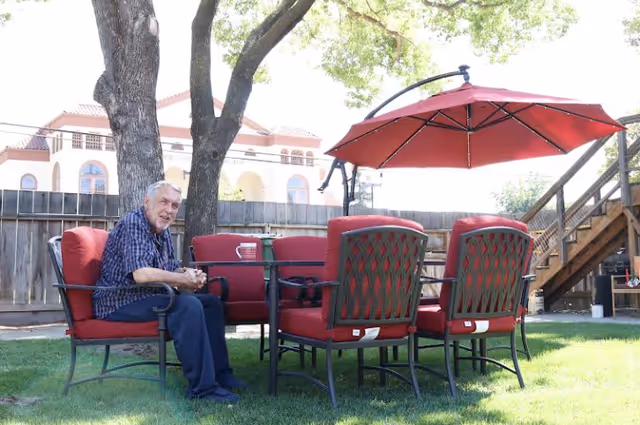 An older man sits at a red-cushioned outdoor patio table with matching chairs and a large red umbrella on a grassy backyard near a wooden fence.