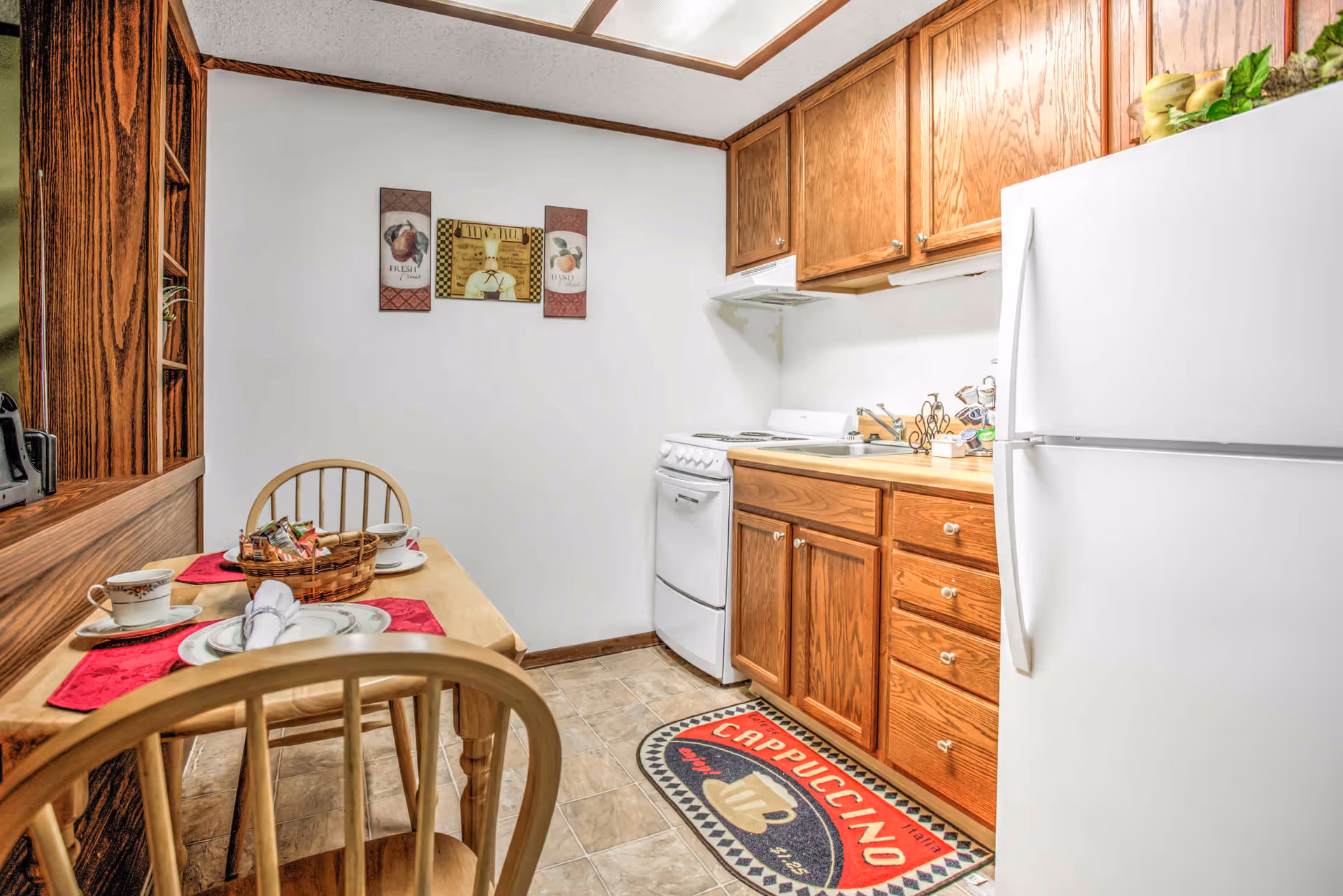 Small kitchen area with wooden cabinets, a white refrigerator, and a white stove. A wooden table with two chairs is set with cups, plates, and a basket of snacks. The wall has three decorative framed pictures, and a colorful rug with the word 'Cappuccino' is on the tiled floor.