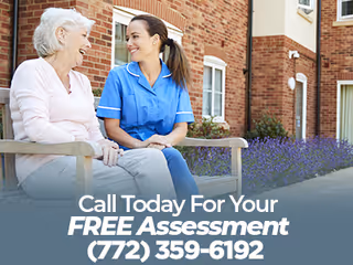 An elderly woman and a caregiver in blue scrubs sitting on a bench outside a brick building, smiling and engaging in conversation with purple flowers and greenery in the background.