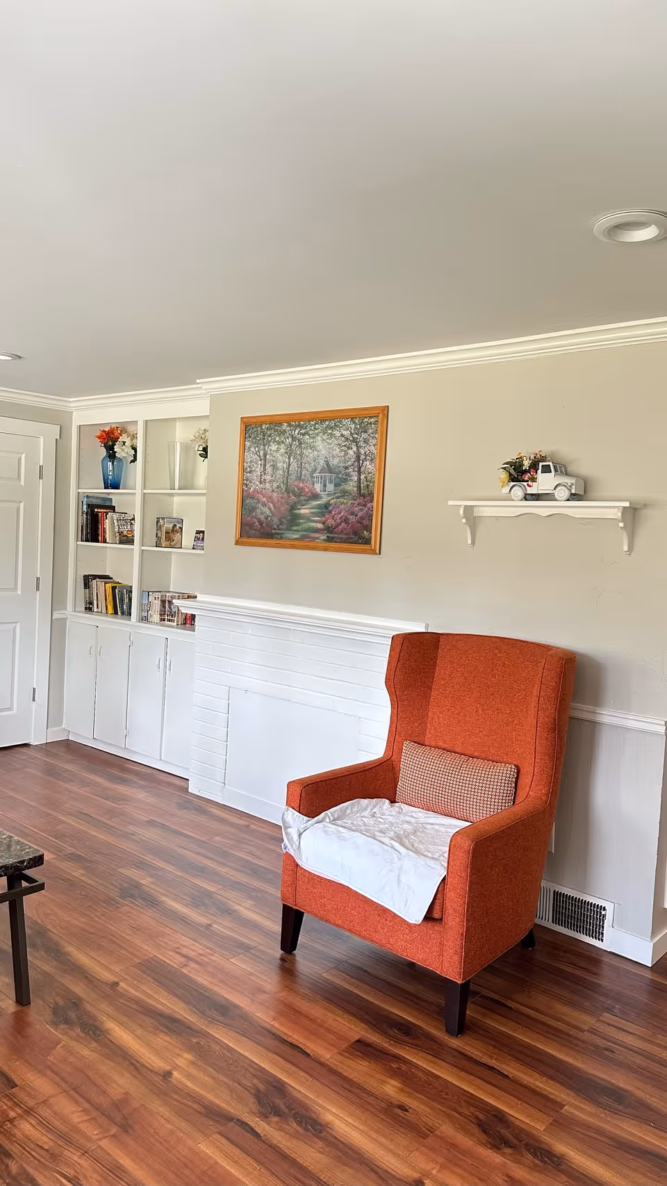 A cozy living room with an orange armchair, built-in white shelving, a framed painting on the wall, and hardwood floors.