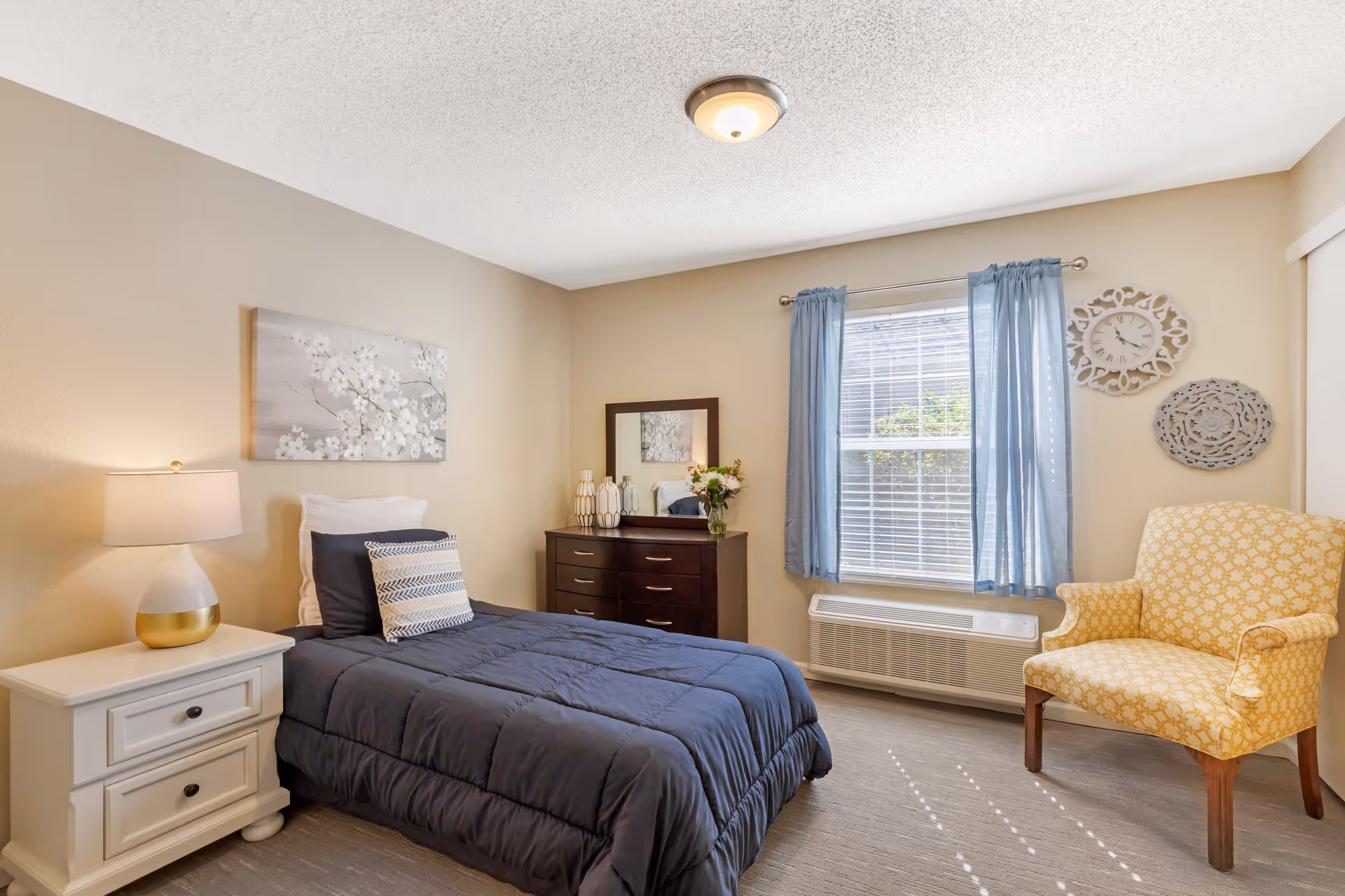 Furnished bedroom with a single bed, white nightstand and lamp, dark dresser with mirror, window with blue curtains, and a yellow patterned armchair.