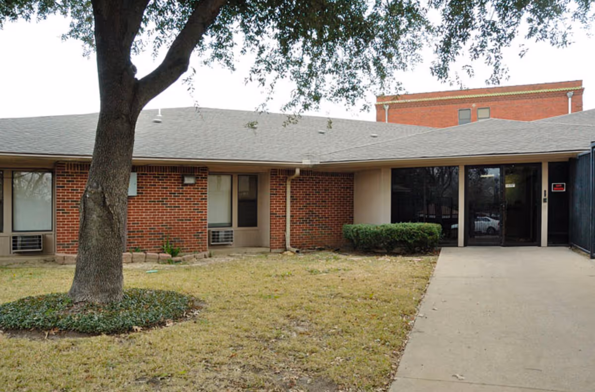 Exterior view of a single-story brick building with a gray roof, a large tree in the front yard, some bushes near the entrance, and a concrete walkway leading to glass doors.