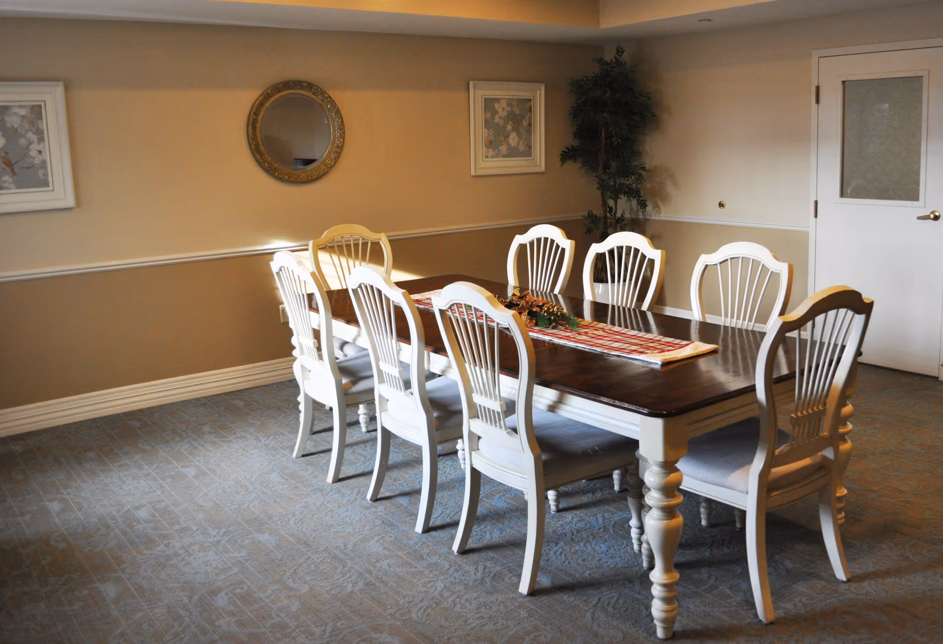 A dining room with a long wooden table and eight white chairs arranged around it. The table has a decorative runner and centerpiece. The room features beige walls with white trim, a round mirror, framed artwork, a potted plant in the corner, and a white door with a frosted glass panel.