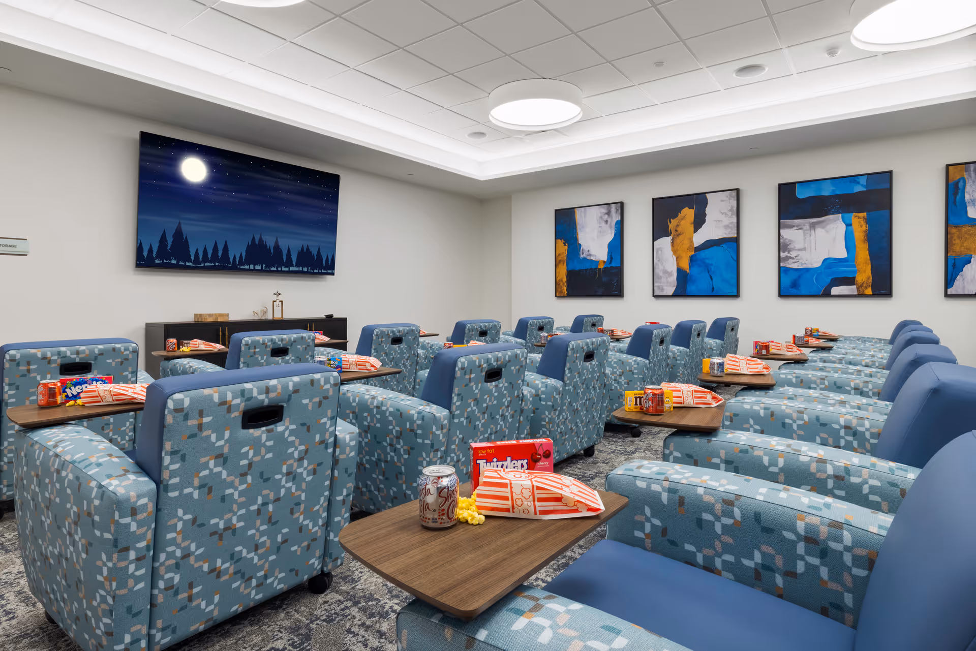 A cozy media room with multiple rows of blue patterned reclining chairs, each equipped with a small wooden table holding popcorn, candy, and soda. A large flat-screen TV on the wall displays a night scene with a full moon and trees. The room has modern ceiling lights and abstract artwork with blue, white, and gold colors on the walls.